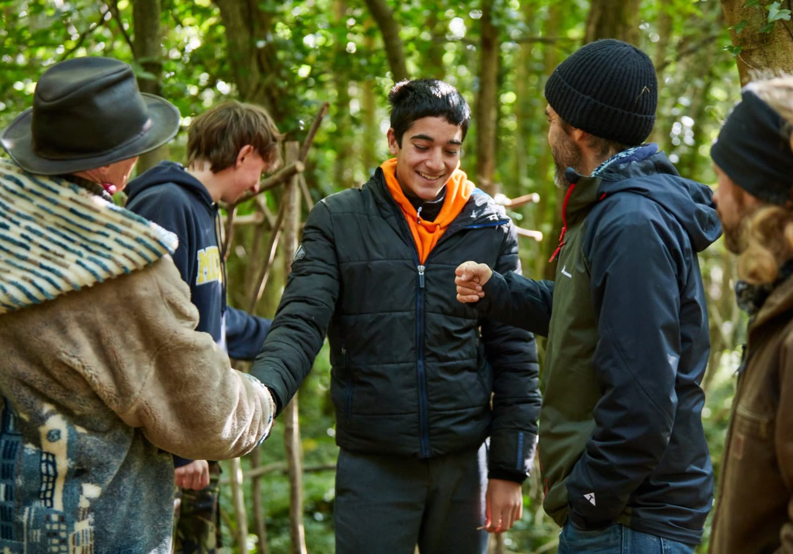 A boy and a mentor bumping fists in the woods, other boys and men around them