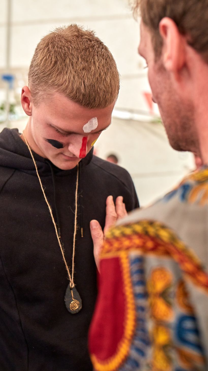 A boy with face paint and a carved pendant, a mentor's hand on his chest