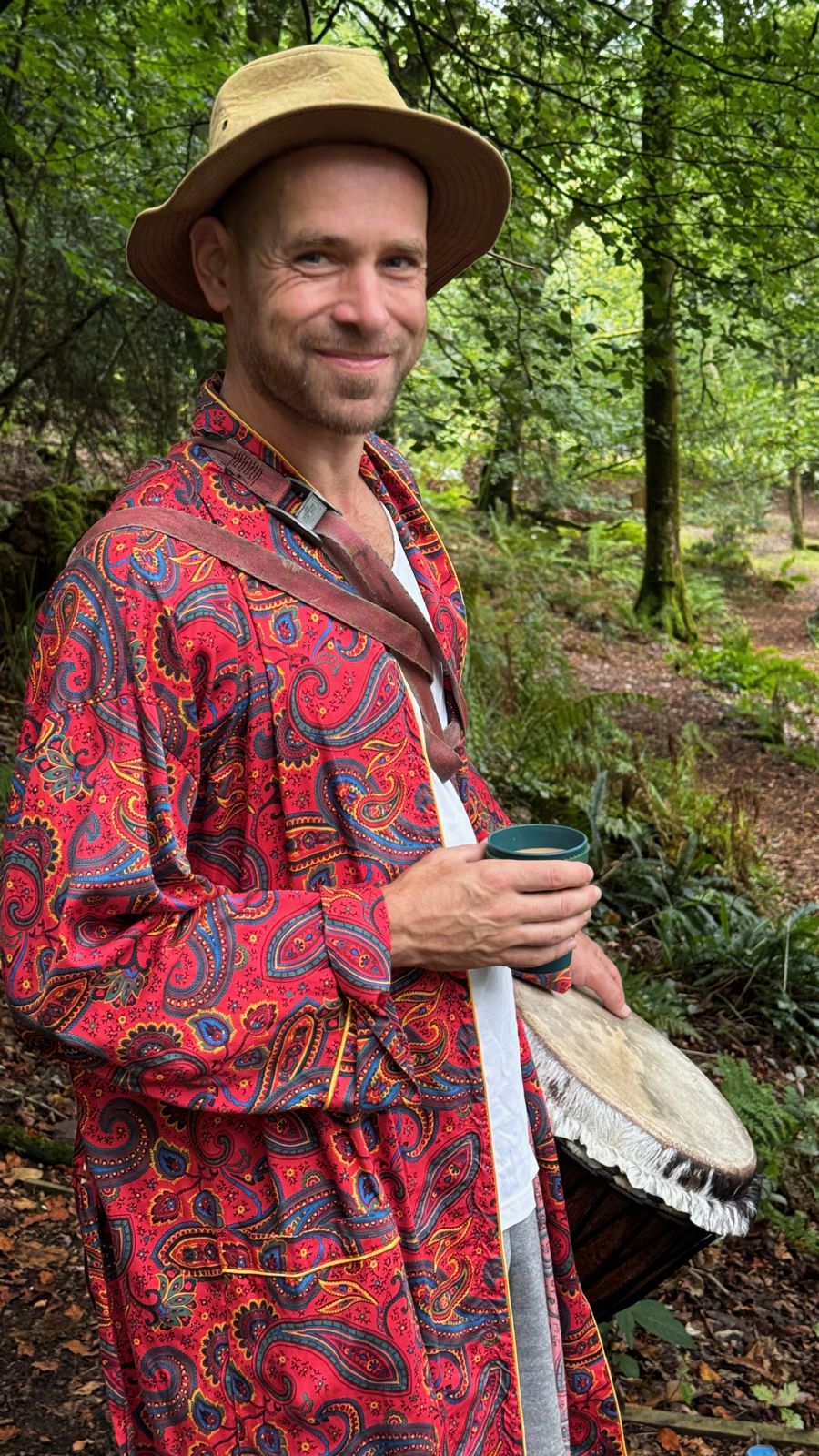 A smiling mentor in a hat and red paisley robe holding a drum in the woods